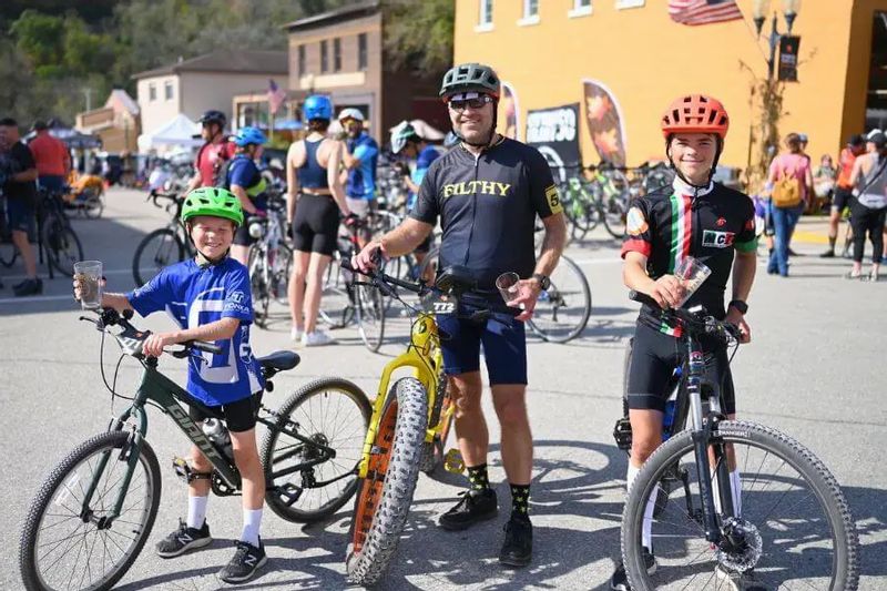 Three Filthy 50 riders of different ages pose with their bikes downtown in Lanesboro, wearing helmets and holding post-race drinks amid a crowd of cyclists.