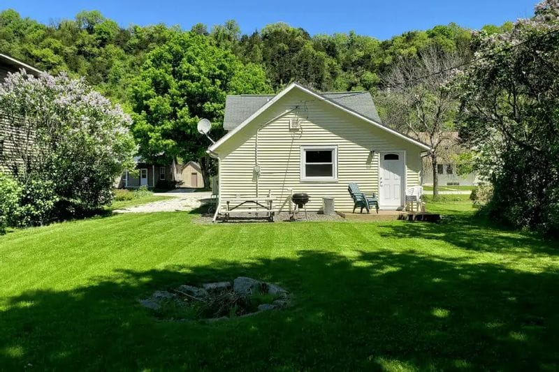 The Cozy Cottage is a small, light-colored vacation rental set on a grassy lawn with outdoor seating, backed by wooded bluffs and trees on a sunny day in Lanesboro.