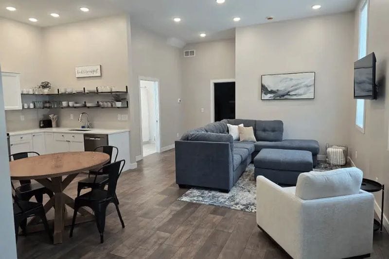 A bright living area inside Parkway Place features an open-concept layout with a modern kitchen, dining table, and comfortable seating area, including a sectional sofa and wall-mounted TV, designed for extended stays in downtown Lanesboro.