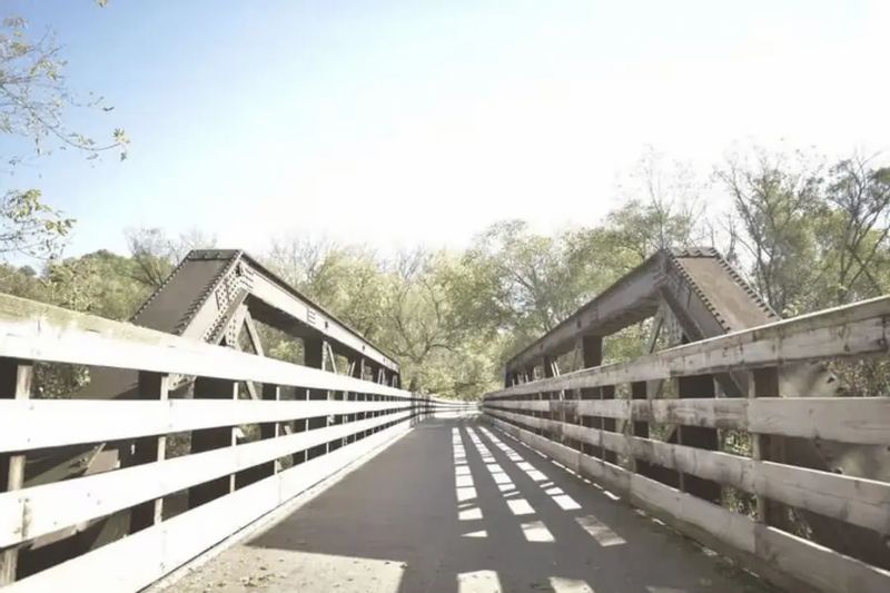 A pedestrian and bike bridge along the Root River State Trail leads through trees near downtown Lanesboro, with sunlight casting long shadows across the wooden railings and paved path.