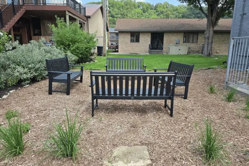An outdoor seating area at Parkway Place features black benches arranged on a landscaped patio, surrounded by greenery and nearby buildings, offering a quiet spot for guests to relax outdoors in downtown Lanesboro.