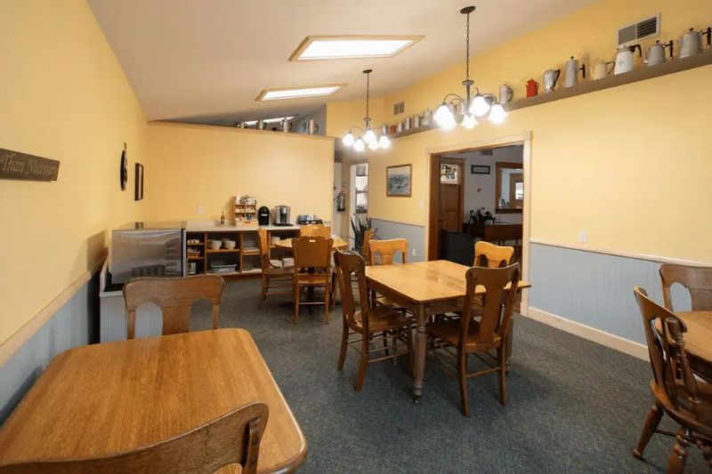 A bright dining and breakfast area at the Cottage House Inn features wooden tables and chairs, soft yellow walls, pendant lighting, and a self-serve kitchen space for guests.