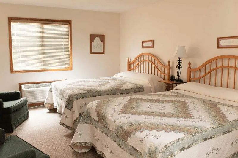 A comfortable guest room at the Cottage House Inn features two beds with coordinated quilts, wooden headboards, soft lighting, and a bright window, creating a simple and relaxing space for overnight stays.