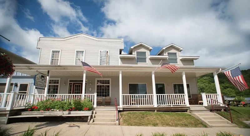 The Cottage House Inn features a historic white clapboard exterior with a wide front porch, rocking chairs, and American flags, welcoming guests along a quiet street in Lanesboro, Minnesota.