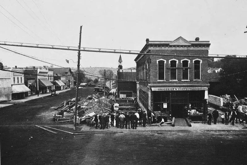 Historic black-and-white photo of a downtown Lanesboro street scene showing a brick commercial building labeled “Groceries & Confectionery” being moved or repositioned, with workers, onlookers, and construction debris gathered around the site.