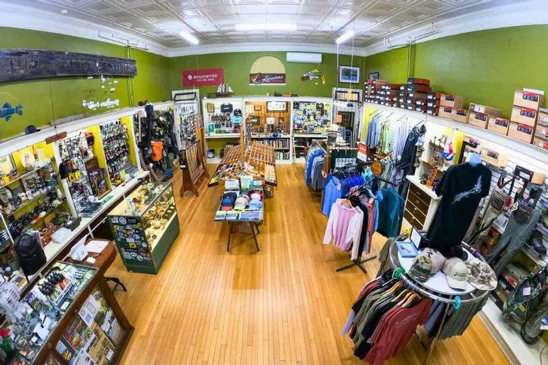 Wide-angle view of the interior of Root River Rod Co. in downtown Lanesboro, showing fishing gear, outdoor apparel, fly rods, books, and accessories displayed on shelves and racks in a bright, historic storefront.