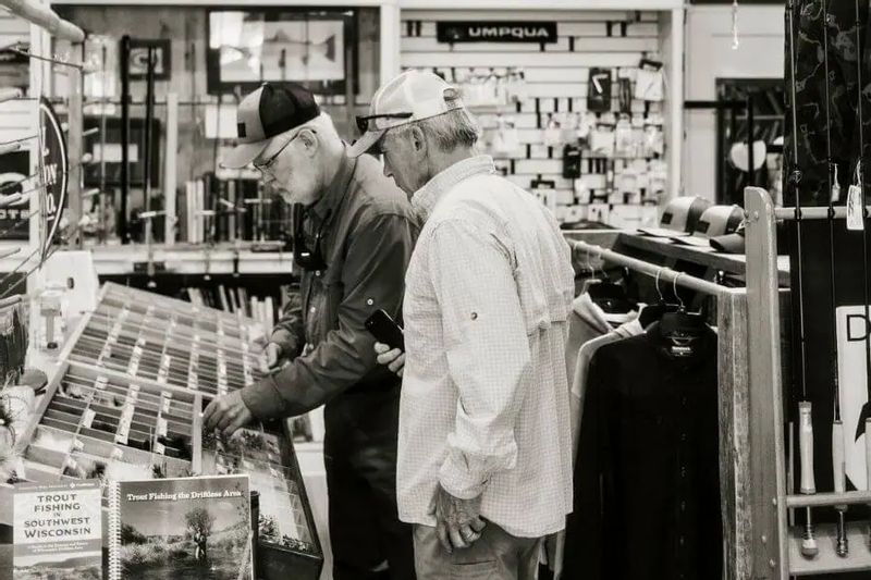 Two men browse fly-fishing gear and tackle inside Root River Rod Co. in Lanesboro, examining flies and equipment displays in a well-stocked local outfitter shop.