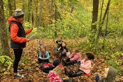 An educator leads a small group of preschool children sitting on fallen leaves in a wooded area, engaging in an outdoor lesson as part of a nature-based preschool program in autumn.
