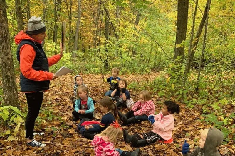 An educator leads a small group of preschool children sitting on fallen leaves in a wooded area, engaging in an outdoor lesson as part of a nature-based preschool program in autumn.