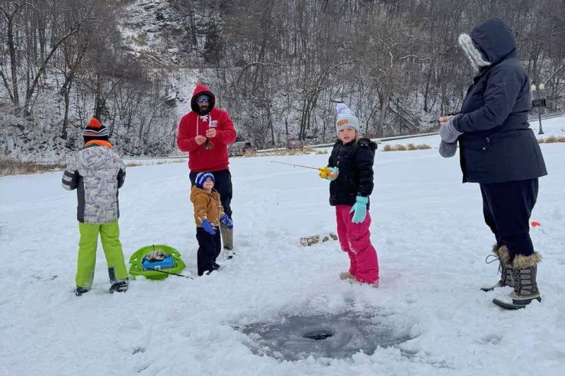 A family with children ice fishing together at the Lanesboro Bass Pond, gathered around a drilled ice hole on the frozen, snow-covered pond.