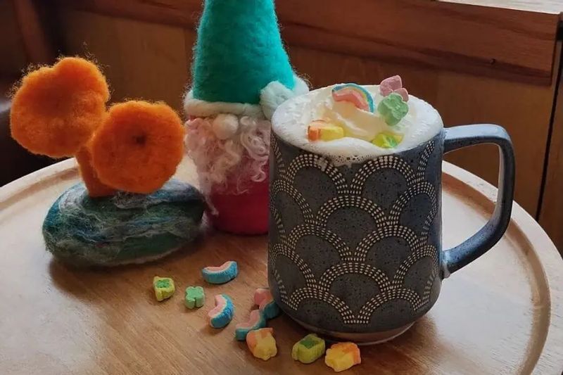 A warm mug of hot cocoa topped with whipped cream and colorful marshmallows sits on a wooden tray beside felted handmade decorations near a window.